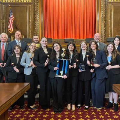 Group of students and mentors in a courtroom posing with trophies and gavels after a mock trial victory.