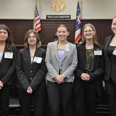 Students and mentors standing in a courtroom setting in front of the judge’s bench during a mock trial event.