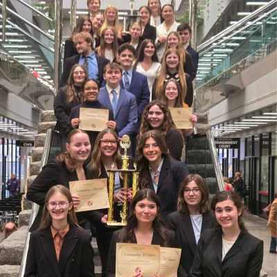 Group of students in business attire standing on stairs holding trophies and certificates at an academic award ceremony.